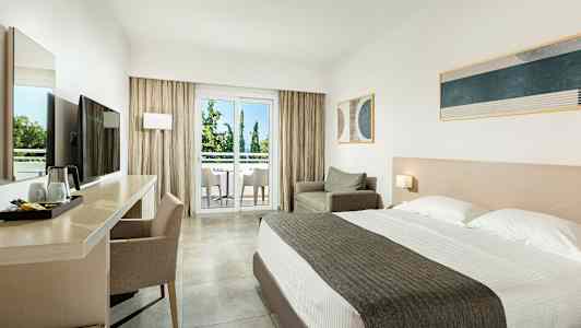 Ett hotellrum med dubbelsäng, skrivbord och stol, fåtölj samt en balkong med utsikt mot träd. (AI-genererad text)