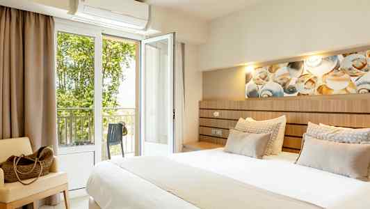 Ett hotellrum med en stor säng, sittmöbel, och öppen balkongdörr med utsikt mot grönska. (AI-genererad text)