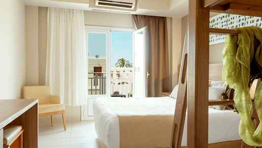 Ett hotellrum med en säng, ett skrivbord och en stol, samt ett öppet glasfönster som vetter mot en balkong. (AI-genererad text)