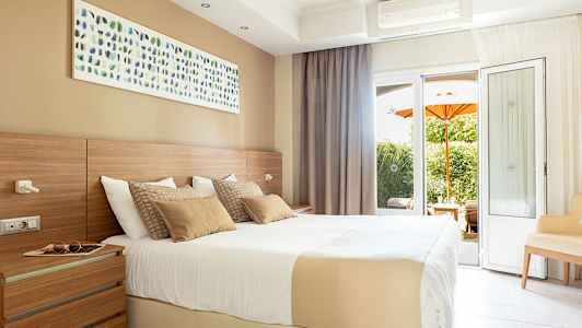 Ett ljust hotellrum med en stor säng, väggkonst och altandörrar med utsikt över en trädgård med en orange parasoll. (AI-genererad text)