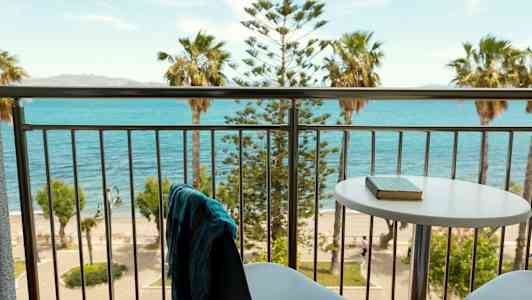 Utsikt från en balkong med bord och stolar mot havet och palmer. (AI-genererad text)