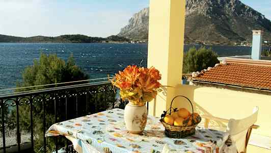 En utsikt från en balkong mot havet, med ett bord dekorerat med en vas med orange blommor och en korg med citroner. (AI-genererad text)