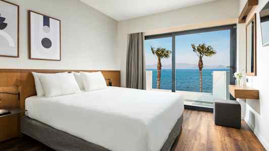 Hotellrum med vit säng och utsikt över havet genom en glasdörr. (AI-genererad text)