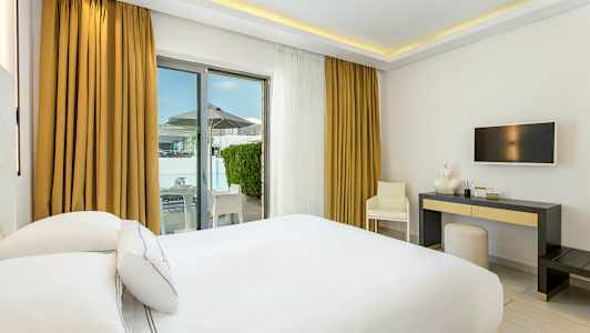 Hotelrum med vit inredning, en dubbelsäng, skrivbord och utgång till terrass. (AI-genererad text)