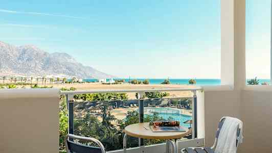 Utsikt från en balkong med bord och stolar mot en pool, landskap med berg samt havet i bakgrunden. (AI-genererad text)