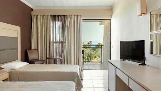 Hotellrum med två sängar, TV, bord och stolar samt en balkong med utsikt över grönska och hav. (AI-genererad text)