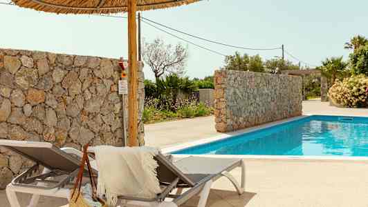 Två solstolar med en handduk och stråväska bredvid en pool. Här finns också en stenmur och palmer i bakgrunden. (AI-genererad text)