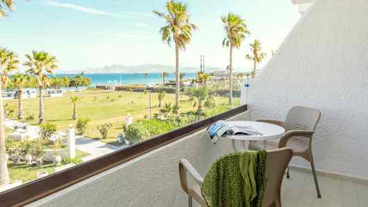En balkong med bord, stolar och utsikt över en grön trädgård med palmer samt havet i bakgrunden. (AI-genererad text)