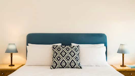 En säng med ett blått huvudgavel, vita kuddar och en dekorativ kudde i mitten. Två nattduksbord med lampor på varje sida. (AI-genererad text)