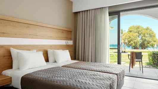 Två bäddar med vita kuddar och grå överkast, vid sida av en glasdörr som leder mot en trädgård med utsikt över havet. (AI-genererad text)