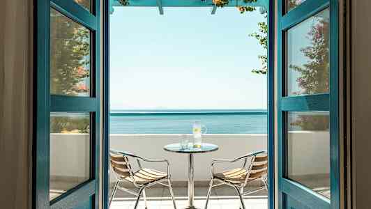 Utsikt genom öppna dörrar mot en terrass med havsutsikt, inklusive ett bord och två stolar. (AI-genererad text)