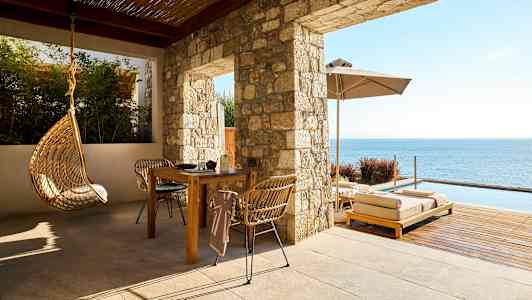 En uteplats med bord, stolar, hängande stol och solsängar vid havet. (AI-genererad text)