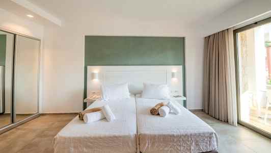 Två enkelsängar med vita sängkläder och hoprullade handdukar, omgivna i ett ljust rum med stor glasdörr. (AI-genererad text)