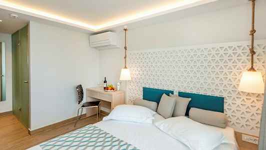 Bilden visar ett modernt hotellrum med en dubbelbädd, vita väggar och en dekorativ panel bakom sängen. Ett skrivbord med stol och frukt syns i rummet. (AI-genererad text)