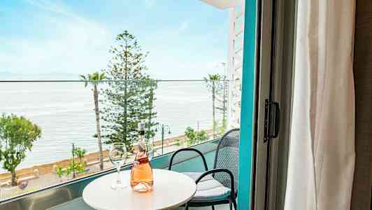 Utsikt från balkong med bord, stolar och en flaska rosévin, mot hav och träd. (AI-genererad text)