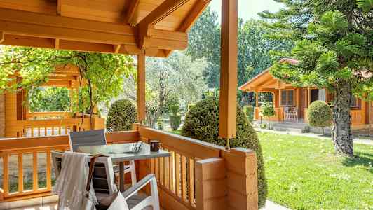 En träveranda med bord och stolar, omgiven av grönska och trähus. (AI-genererad text)