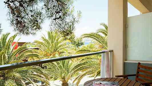 Utsikt från en balkong med träbord och stol, mot palmer och hav i bakgrunden. (AI-genererad text)