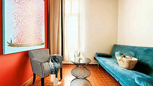 Ett vardagsrum med en blå soffa, en mönstrad stol och ett runt bord, samt en stor färgglad tavla på väggen. (AI-genererad text)