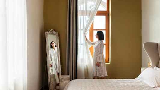 En kvinna står vid ett fönster och håller i gardinen, med en stor spegel bredvid. (AI-genererad text)