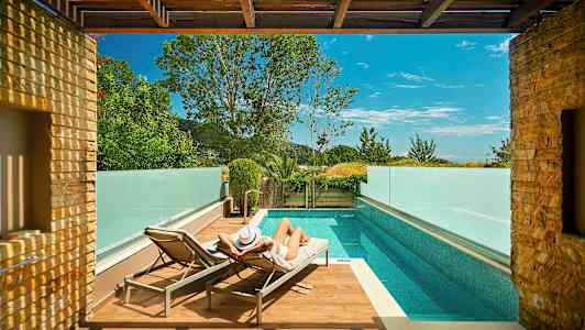 En person ligger på en solstol vid en pool omgiven av glasräcken och träd. (AI-genererad text)