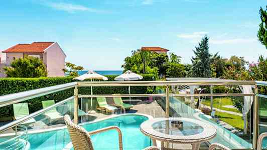 Terrass med bord och stolar som vetter mot en utomhuspool, grönska och hav i bakgrunden. (AI-genererad text)