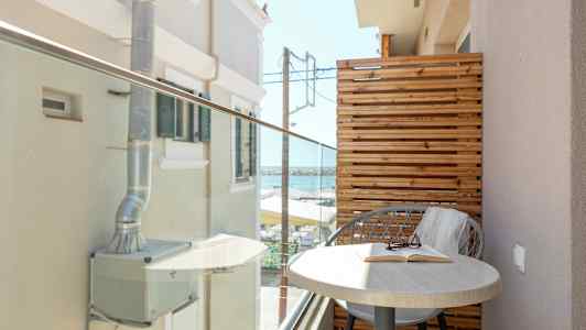 En liten balkong med ett bord och en stol, samt utsikt mot byggnader och havet. (AI-genererad text)