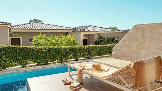 En solstol vid en pool med en stråhatt och en handduk på. I bakgrunden syns bostadshus och vegetation. (AI-genererad text)