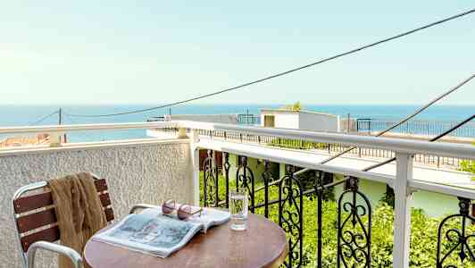 En balkong med utsikt över havet, ett bord med ett glas vatten, en tidning och solglasögon. (AI-genererad text)