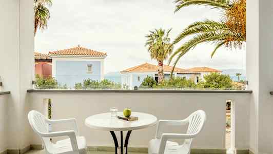 Två vita plaststolar och ett bord på en balkong med utsikt över byggnaden med röda tak och palmer. (AI-genererad text)