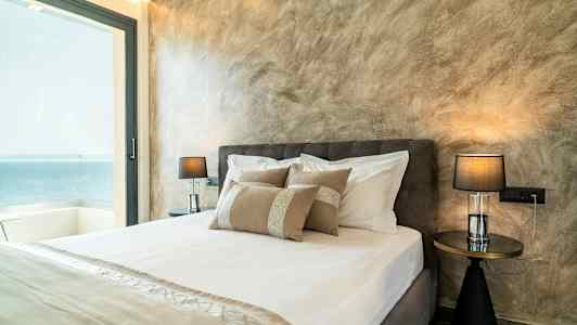 Ett hotellrum med en dubbelsäng, dekorativa kuddar och nattduksbord med lampor, samt havsutsikt genom en stor glasdörr. (AI-genererad text)