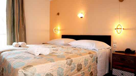 Ett sovrum med två enkelsängar täckta med ett blommigt överkast, vita kuddar och vikta handdukar, samt nattlampor på väggen. (AI-genererad text)