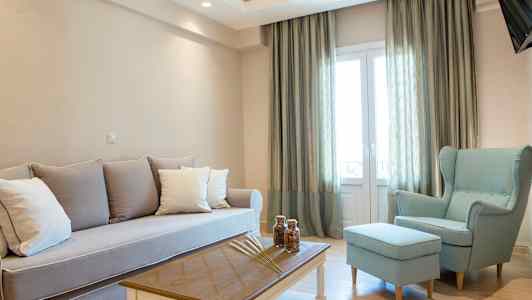 Ett vardagsrum med ljusa möbler, inklusive en soffa, en fåtölj och ett soffbord, bredvid stora fönster med gardiner. (AI-genererad text)