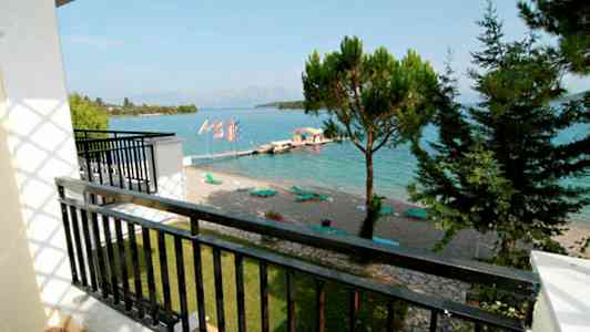 Utsikt från en balkong mot en strand med solstolar vid en klarblå havsvik. Träd och moln i bakgrunden. (AI-genererad text)