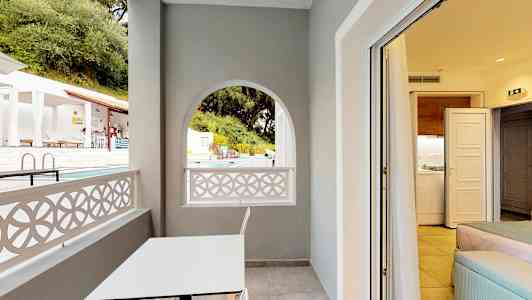 En balkong med ett vitt bord och en stol, utsikt mot poolområdet, och insyn till ett rum. (AI-genererad text)