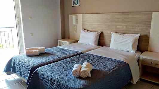 Rummet har två enkelsängar med blå överkast och vita kuddar, samt hoprullade handdukar placerade på sängen. (AI-genererad text)