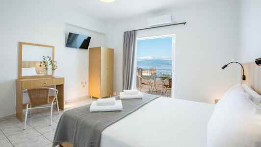 Ett ljust hotellrum med en dubbelsäng, skrivbord, spegel och en balkong med utsikt över havet. (AI-genererad text)