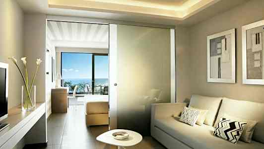 Ljust hotellrum med soffa, låg bord och en glasvägg mot sovrumsdel med dubbelsäng. Balkong med havsutsikt syns. (AI-genererad text)