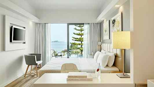 Ett hotellrum med modernt inredning och fönster som vetter mot en balkong med utsikt över havet. (AI-genererad text)