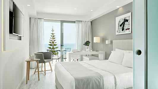 Modernt hotellrum med dubbelsäng, skrivbord och skjutdörr ut till en balkong med havsutsikt. (AI-genererad text)