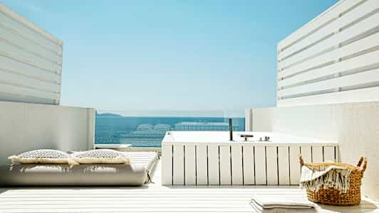 En vit terrass med en badtunna, kuddar och utsikt över havet. (AI-genererad text)