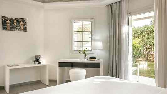 Ett ljust hotellrum med ett skrivbord och en fönsterdörr ut mot en trädgård. (AI-genererad text)