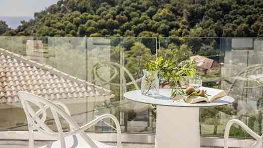 Ett bord med glasvas, frukt, bok och glas på en terrass med utsikt över träd och tak. (AI-genererad text)