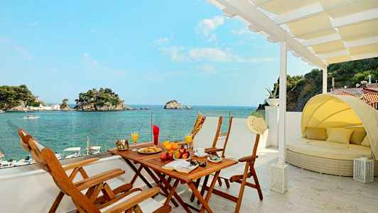 Utsikt från en terrass med matbord och solsäng mot havet och små öar. (AI-genererad text)
