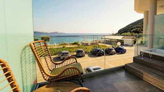 Utsikt från balkong med rottingstolar och bord, glasräcke, badkar på en plattform samt bilar parkerade vid stranden med havet i bakgrunden. (AI-genererad text)