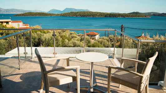 Två stolar och ett mindre bord på en terrass med glasräcke, utsikt mot havet och skogklädda öar. (AI-genererad text)