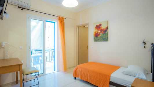 Ett enkelrum med en säng klädd i en orange filt, skrivbord och stol, samt en glasdörr som leder till en balkong. (AI-genererad text)