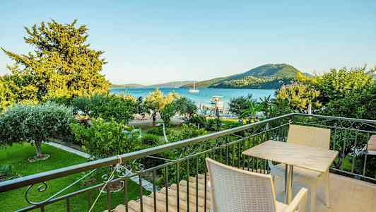 En balkong med bord och stolar, utsikt över en grön trädgård, havet och berg i bakgrunden. (AI-genererad text)