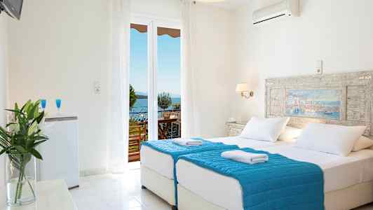 Ett ljust hotellrum med dubbelsäng och blå sängklädsel. Det finns en balkong med utsikt över havet. (AI-genererad text)