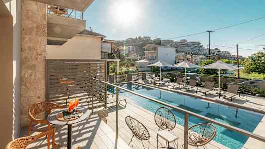 Bild av en solig balkong med bord och stolar, en poolområde nedanför med solstolar och parasoller. (AI-genererad text)