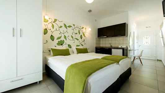 Ett hotellrum med en dubbelsäng täckt med vit sängkläder och gröna filtar, en garderob, ett pentry och en dörr i bakgrunden. (AI-genererad text)